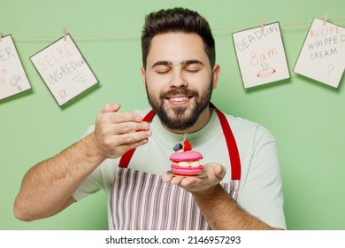 Young Happy Male Chef Confectioner Baker Man 20s In Striped Apron Hold Sniff Scent Of Pink Cake Muffin Macaroon Isolated On Plain Pastel Light Green Background Studio Portrait. Cooking Food Concept