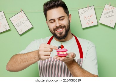 Young Happy Male Chef Confectioner Baker Man 20s In Striped Apron Adorn Decorate Little Pink Cake Muffin Macaroon Isolated On Plain Pastel Light Green Background Studio Portrait. Cooking Food Concept