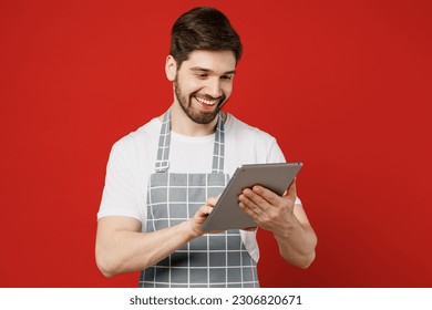 Young happy male housewife housekeeper chef cook baker man wears grey apron hold use digital tablet pc computer browsing internet isolated on plain red color background studio. Cooking food concept - Powered by Shutterstock