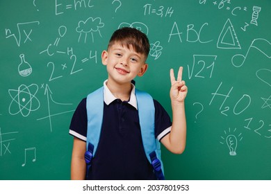 Young happy male kid school boy 5-6 years old in t-shirt backpack showing victory sign isolated on green wall chalk blackboard background studio. Childhood children kids education lifestyle concept.