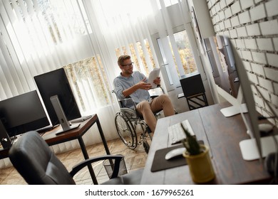 Young Happy Male Office Worker In A Wheelchair Reading Some Documents And Smiling While Sitting Near Printer At His Workplace In The Bright Modern Office. Disability And Handicap Concept