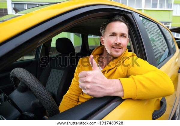 Young happy male taxi driver sits behind the wheel\
of a taxi and shows like