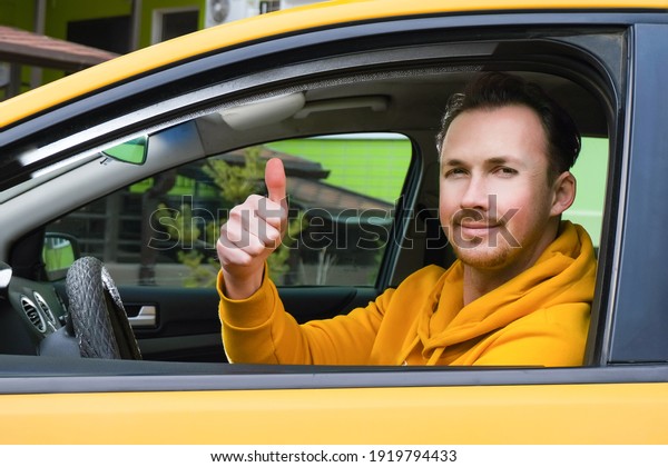 Young happy male taxi driver sits behind the wheel\
of a taxi and shows like
