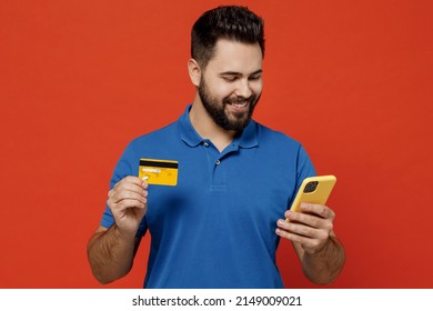 Young Happy Man 20s In Basic Blue T-shirt Using Mobile Cell Phone Hold Credit Bank Card Doing Online Shopping Order Delivery Booking Tour Isolated On Plain Orange Background. People Lifestyle Concept