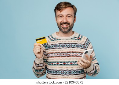 Young Happy Man 30s He Wear Sweater Using Mobile Cell Phone Hold Credit Bank Card Doing Online Shopping Order Delivery Booking Tour Isolated On Plain Pastel Light Blue Cyan Background Studio Portrait