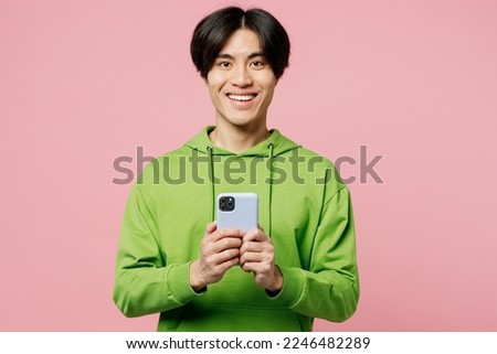Young happy man of Asian ethnicity wear green hoody look camera hold in hand use mobile cell phone in blue case isolated on plain pastel light pink background studio portrait. People lifestyle concept