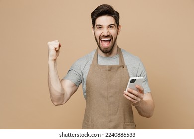 Young happy man barista barman employee wear brown apron work in coffee shop hold use mobile cell phone do winner gesture isolated on plain pastel light beige background Small business startup concept - Shutterstock ID 2240886533