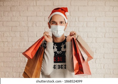 Young Happy Man In Christmas Sweater, Medical Face Mask Hold Shopping Bags And Smile. Cheerful Hipster Man In Santa Hat, Protective Face Mask With Paper Bags In Hands. Winter Holidays Sale In Mall