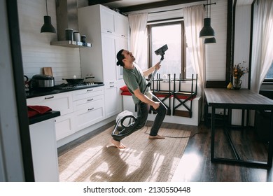 Young Happy Man Doing Cleaning And Dancing With Vacuum Cleaner In Kitchen And Living Room At Home. A Handsome Guy Is Vacuuming A Carpet, Dancing And Singing In A Cozy Room On A Sunny Day