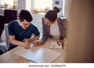 Young happy man with down syndrome sketching on piece of paper with his special education teacher at home. - Shutterstock ID 2139861079