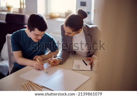 Young happy man with down syndrome sketching on piece of paper with his special education teacher at home.