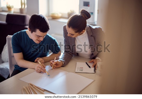 Young happy man with\
down syndrome sketching on piece of paper with his special\
education teacher at\
home.