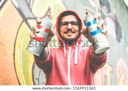 Young happy man drawing with two smiling sprays  - Graffiti artist painting with aerosol color cans on the wall - Urban lifestyle, youth, millennial generation and street art concept - Focus on bottle