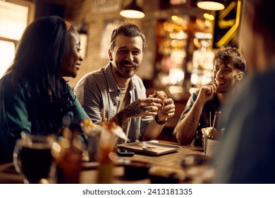 Young happy man eating burger while communicating with his friends in a pub.  - Powered by Shutterstock