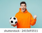 Young happy man fan he wears orange hoody casual clothes do winner gesture cheer up support football sport team hold in hand soccer ball watch tv live stream isolated on plain blue color background