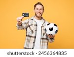 Young happy man fan wear brown shirt casual clothes cheer up support football sport team hold in hand soccer ball watch tv live stream hold in hand credit bank card isolated on plain yellow background