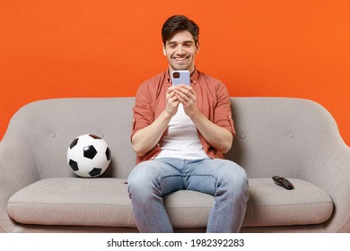 Young happy man football fan wear shirt support team with soccer ball sitting on sofa home watch tv live stream using mobile cell phone makes bets isolated on orange background. People sport concept.
