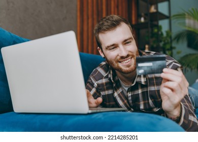 Young Happy Man He Wears Brown Shirt Laptop Pc Computer Hold Credit Bank Card Shopping Online Order Delivery Book Tour Sit On Blue Sofa In Own Living Room Apartment Stay Home Indoor Flat On Weekends.