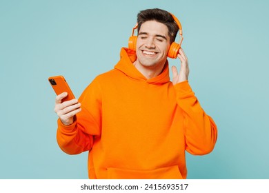 Young happy man he wears orange hoody casual clothes listen to music in headphones use mobile cell phone isolated on plain pastel light blue cyan color background studio portrait. Lifestyle concept - Powered by Shutterstock