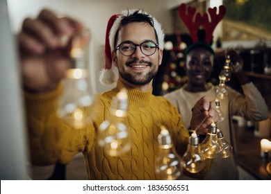 Young happy man and his girlfriend making preparations for Christmas party and decorating their home with glowing garland.