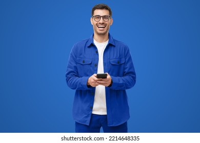 Young happy man laughing as he is looking at camera, holding his phone, isolated on blue background - Powered by Shutterstock