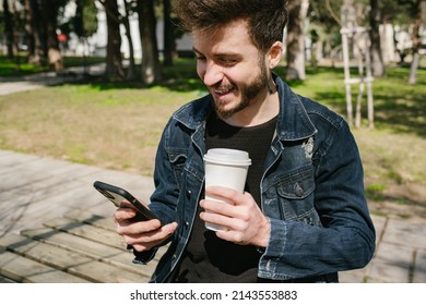 Young Happy Man Looking Phone Screen While Holding A Cup Of Coffee At Outside. Man Breathing Fresh Air At The Outdoor While Drinking Coffee. He Feel Relax And Happy With Healthy Lifestyle.