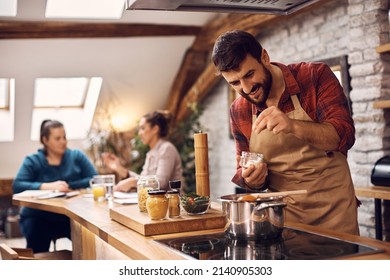 Young happy man preparing food and adding salt into cooking pot in the kitchen. His friends are in the background.  - Shutterstock ID 2140905303