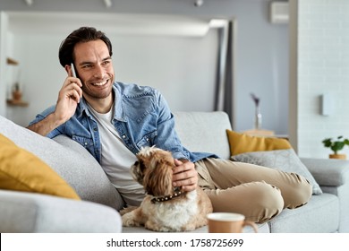 Young Happy Man Relaxing On The Sofa With His Dog While Making A Phone Call Over Cell Phone.