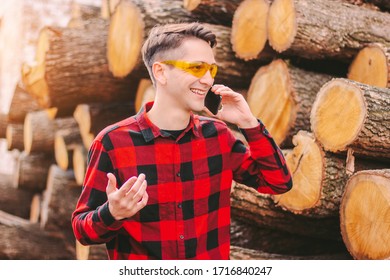 Young Happy Man Sawmill Warehouse Workman In Protective Glasses Talking Mobile Phone And Smiling. Business Man Owner Of Small Wood Material Production Factory Inspecting Cut Tree Logs. Lumber Storage