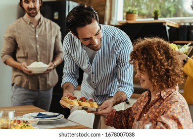 Young Happy Man Serving Food To His Girlfriend While Having Lunch With Friends At Home. 