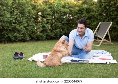 Young Happy Man Sits On Picnic Blanket On The Backyard And Plays With His Cute And Adorable Friend. Man Training His Pet Dog To Give A Paw