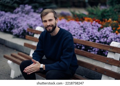 Young Happy Man Sits And Rests On Bench In Park Against Background Of Blooming Flowerbed.