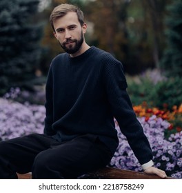 Young Happy Man Sits And Rests On Bench In Park Against Background Of Blooming Flowerbed.