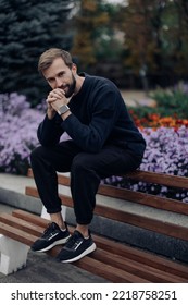 Young Happy Man Sits And Rests On Bench In Park Against Background Of Blooming Flowerbed.