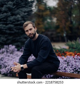 Young Happy Man Sits And Rests On Bench In Park Against Background Of Blooming Flowerbed.