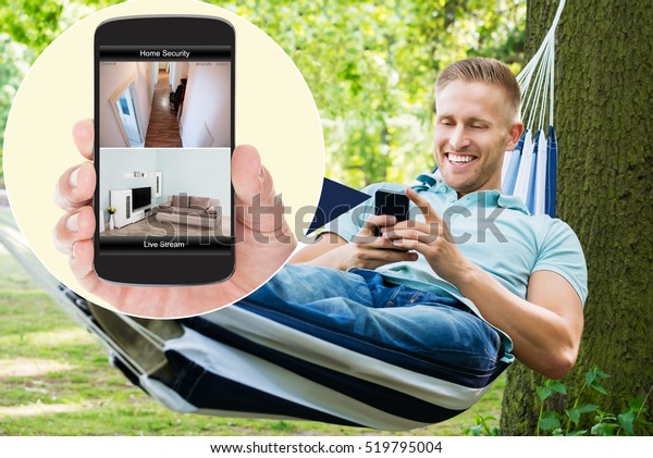 Young Happy Man Sitting On Hammock Looking At\
Home Security System On\
Mobilephone