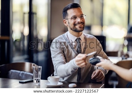 A young happy man in smart casual is sitting in coffee shop and paying with cashless technology with credit card in coffee shop.