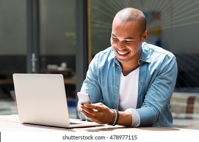 Young Happy Man Smiling And Holding A Smartphone Sitting Outdoor With Laptop. Portrait Of Laughing African Guy Reading A Message With Smartphone In Cafe. Man Smiling And Writing A Phone Message.