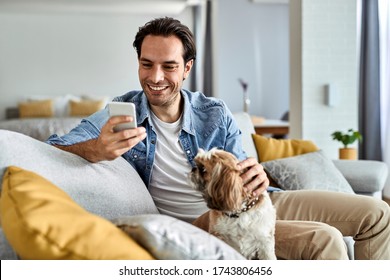 Young Happy Man Text Messaging On Cell Phone While Sitting On The Sofa With His Dog.