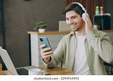 Young happy man wear casual clothes headphones sits alone at table in coffee shop cafe restaurant indoors work or study on laptop pc computer use cell phone. Freelance mobile office business concept