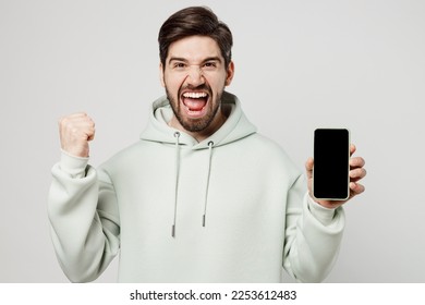 Young happy man wear mint hoody hold in hand use mobile cell phone with blank screen workspace area do winner gesture isolated on plain solid white background studio portrait. People lifestyle concept - Shutterstock ID 2253612483