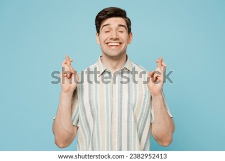 Young happy man wears striped shirt casual clothes waiting for special moment, keeping fingers crossed, making wish, eyes closed isolated on plain pastel light blue cyan background. Lifestyle concept