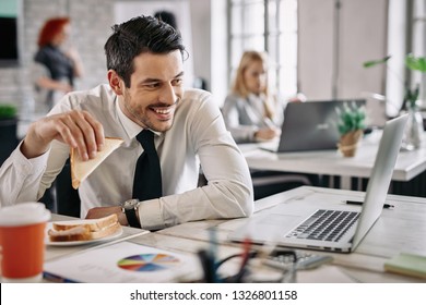 Young happy manager eating sandwich and using computer on lunch break in the office. There are people in the background.  - Powered by Shutterstock