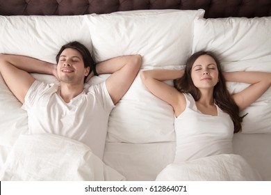 Young Happy married couple wearing white resting with eyes closed in bed, lying with hands behind the head after waking up in the morning, or going to sleep at night. Lovers relaxing in bed. Top view
