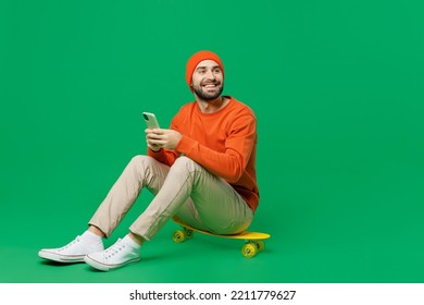 Young happy minded pensive fun cool caucasian man 20s wearing orange sweatshirt hat sit on skateboard hold use mobile cell phone look aside isolated on plain green color background studio portrait. - Powered by Shutterstock