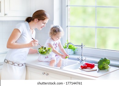 Young Happy Mother And Her Cute Curly Toddler Daughter Washing Vegetables Together In A Kitchen Sink Getting Ready To Cook Salad For Lunch In A Sunny White Kitchen With A Big Garden View Window