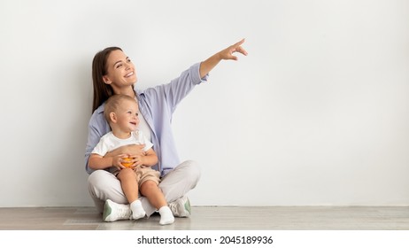 Young Happy Mother With Infant Baby Sitting On Floor Near White Wall And Pointing Aside At Copy Space, Smiling Mom And Child Demonstrating Free Place For Your Design Or Advertisement, Panorama - Powered by Shutterstock