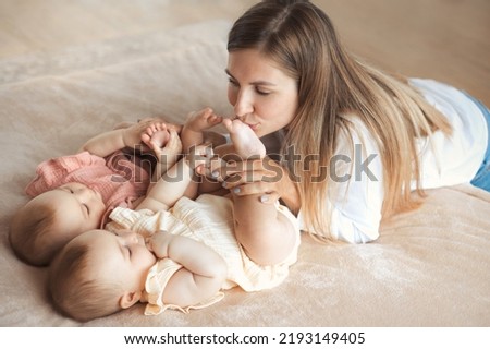 Young happy mother kissing barefoot legs of two baby twin sisters on the soft bed at home