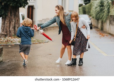 Young Happy Mother With Two Kids, Little Boy And Girl, Playing Outside On A Fresh Rainy Day. Stylish Family Having Fun Outdoors