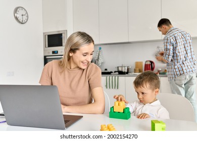 Young Happy Mother Working From Home Office Using Laptop Computer Sitting At Kitchen Table With Cute Kid Child Son At Home While Dad Cooking. Parent And Child Distance Virtual Work In Family Life.